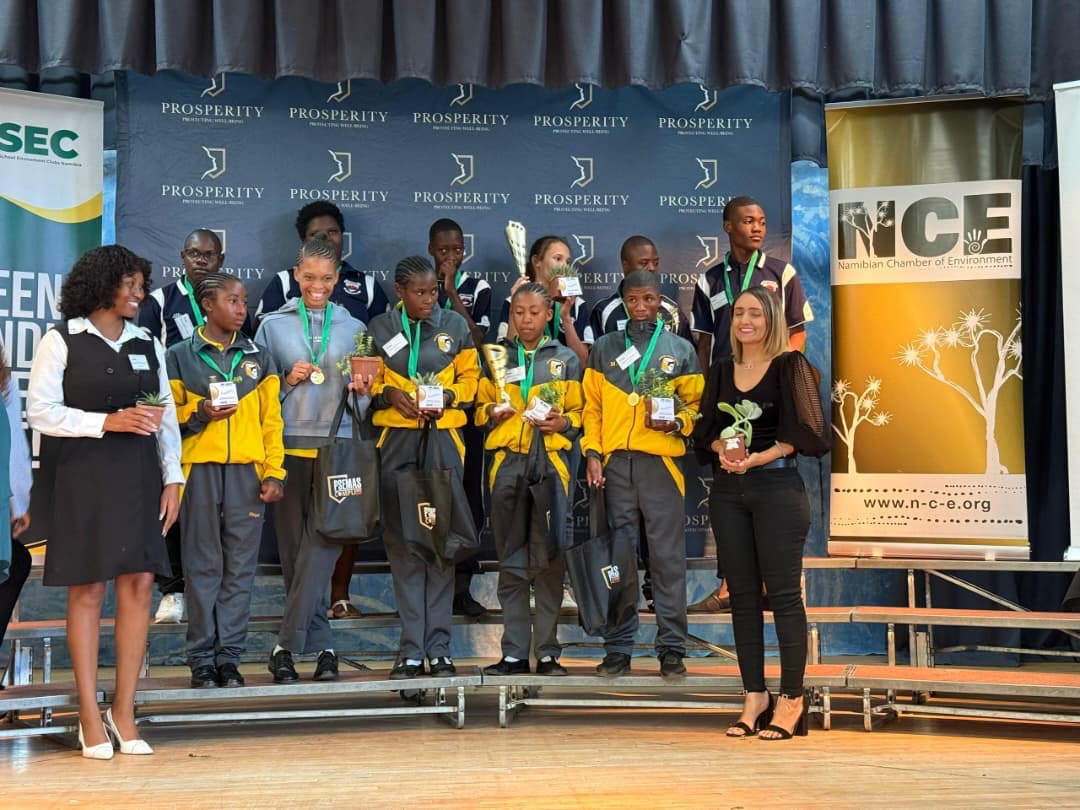 A second school's team receiving medals and trophies on stage with NCE banner
